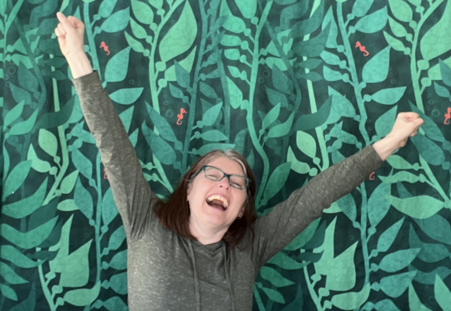 Becka is standing in front of a leafy green curtain making a celebration gesture with her hands in the air.