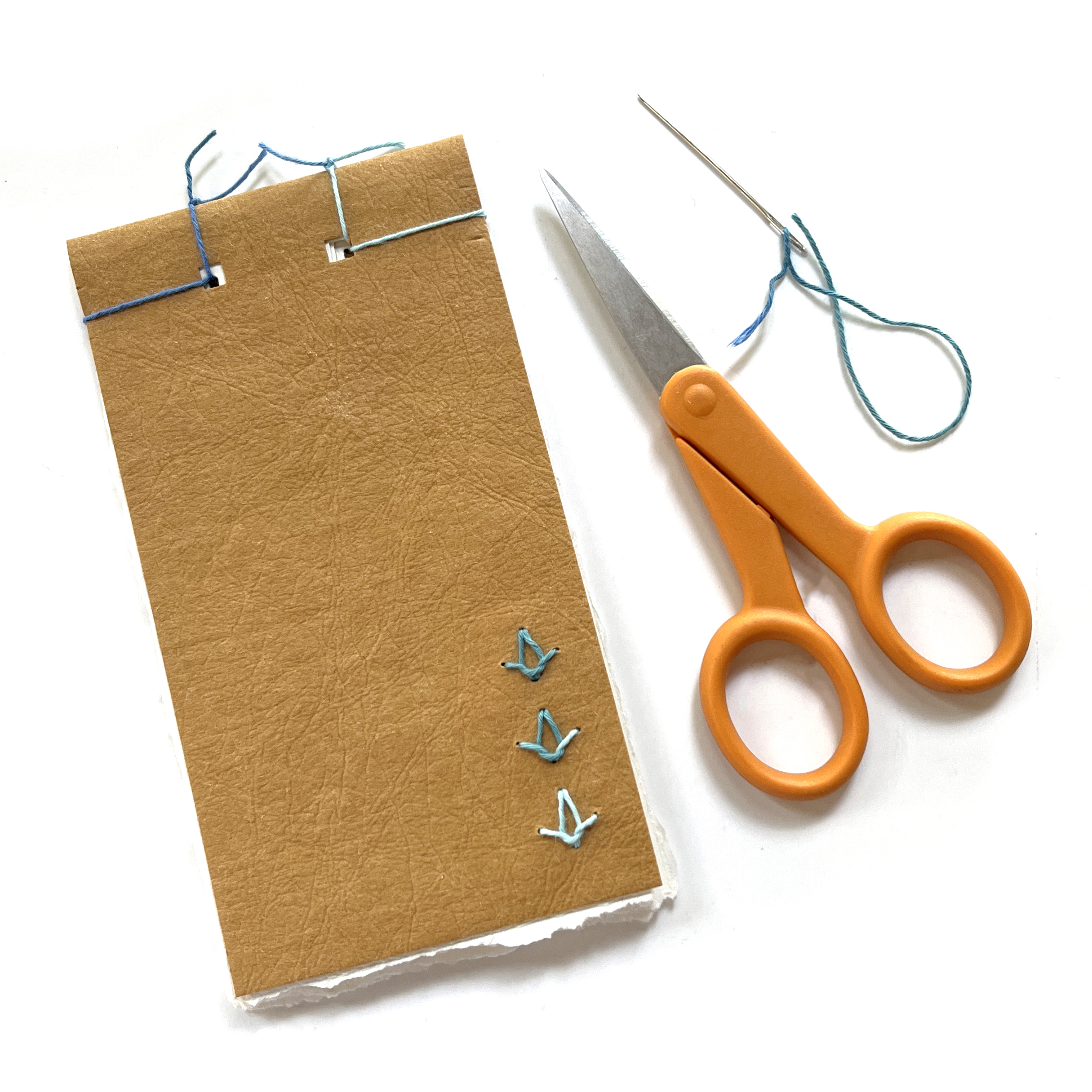 Photo shows a small tan notebook with a pair of scissors and a needle and thread.
